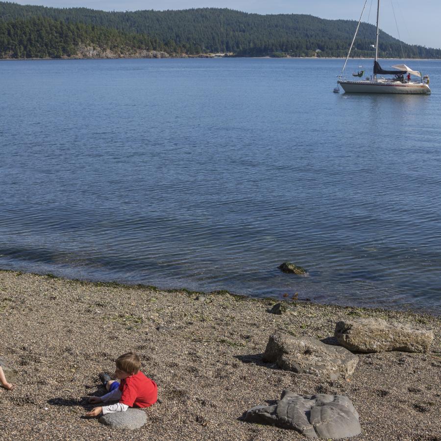 Image of two youths playing on the rocky beach in the foreground. In the midground a sailboat can be seen on the water. The boat is white with a red stripe horizontally across the body. The sails are down. In the background, land across the water is visible with forested hills.