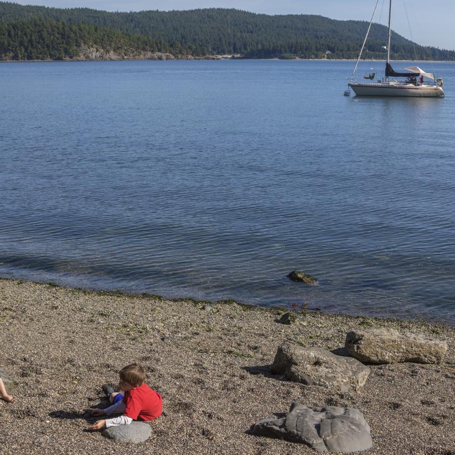 Image of two youths playing on the rocky beach in the foreground. In the midground a sailboat can be seen on the water. The boat is white with a red stripe horizontally across the body. The sails are down. In the background, land across the water is visible with forested hills. 