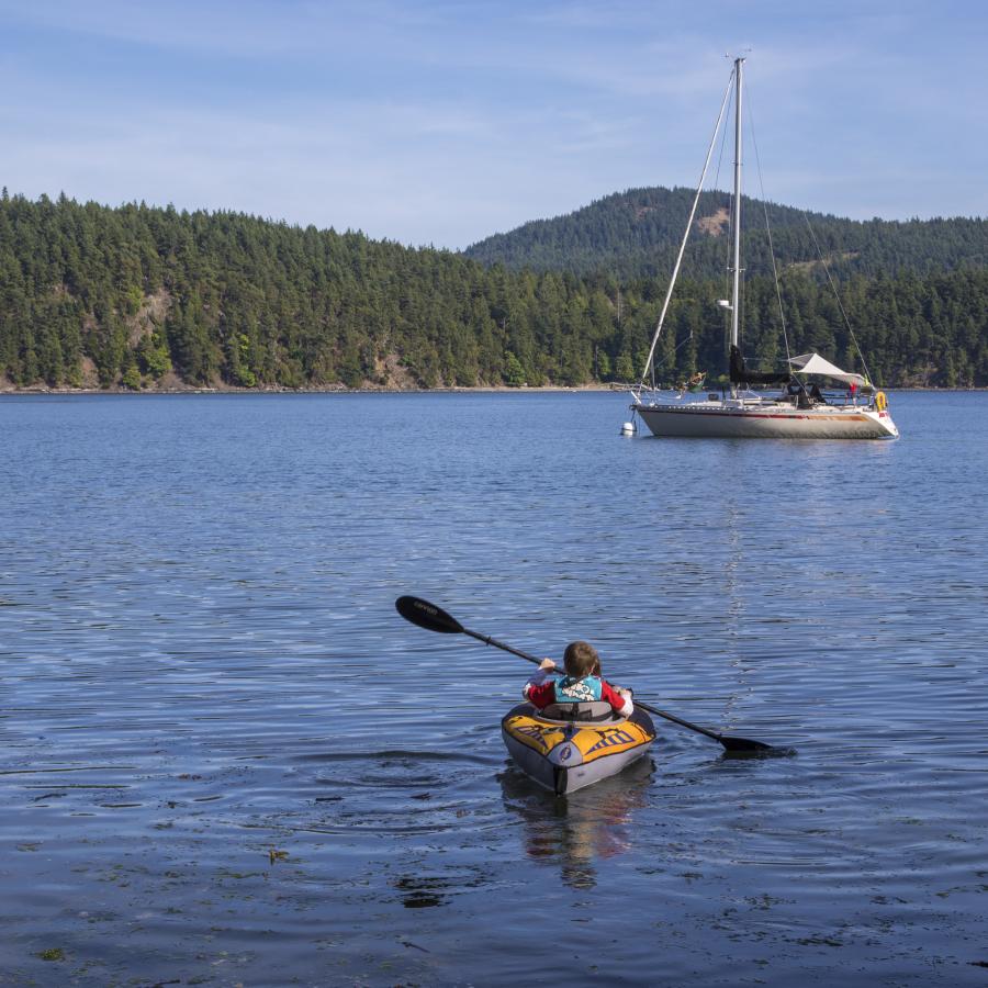 Two youths kayaking in a yellow and grey kayak. There is a sailboat in the midground. The water is bright blue - a similar hue to the sky. In the background you can see the forested hills of the other side of the water.