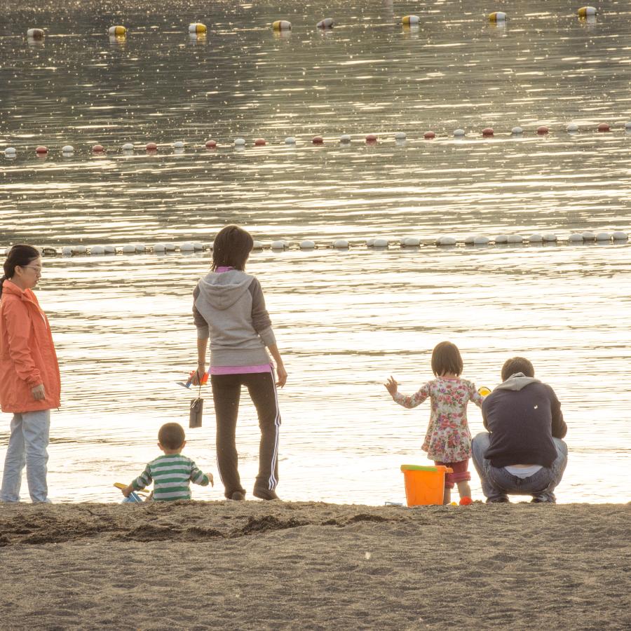 Family with 2 young children and 3 adults exploring the sand at Sunset Beach. One adult is wearing light colored jeans and a peach colored jacket, another is in black track pants and a grey and pink sweatshirt, and the third in a grey or blue sweatshirt and jeans. One of the youth is wearing a green and white stripped shirt and the other is in a floral pattern. The swimming buoys are visible on the water and it appears to be golden hour where the sun creates a soft, golden glow.