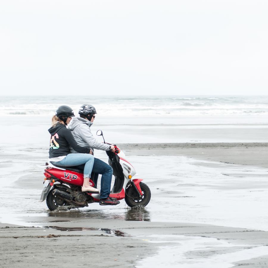 two people riding red scooter on hardpacked sand of the beach by shoreline through receding pools of water