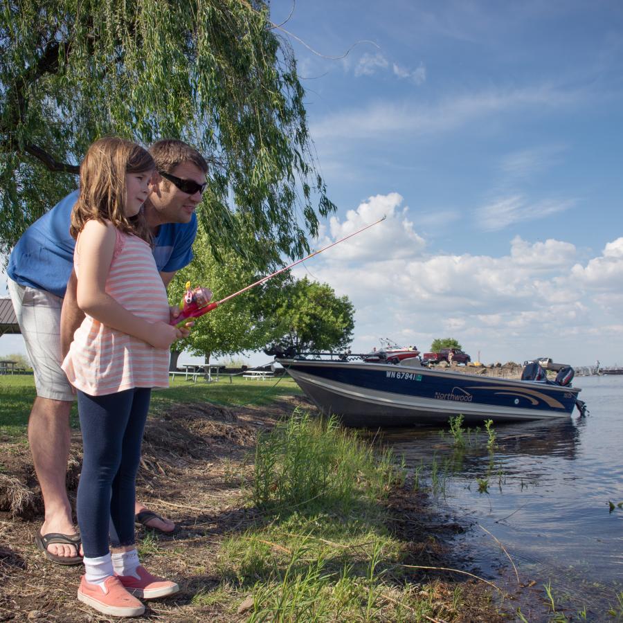 A father, wearing a blue shirt, khaki shorts and sunglasses, teaches his daughter, wearing a pink and white striped shirt with blue pants and pink shoes, how to fish. A beached boat next to the grass sits behind them.