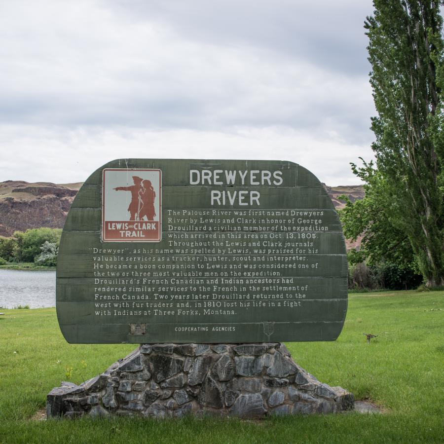 An interpretive sign of the Drewyers River and the Lewis and Clark Trail sits on rocks in a green grassy lawn. In the background, the lawn meets the river with a picnic table and barbeque brazier near the bank. The rocky hillside can be seen behind the sign.