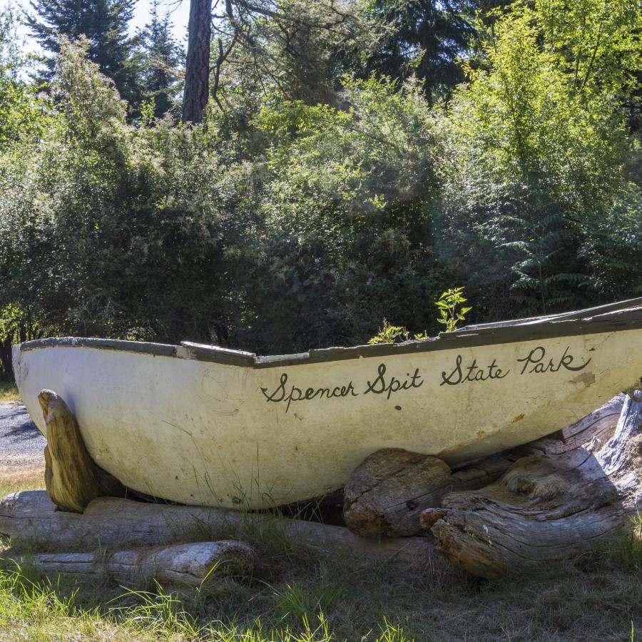 An old cream and brown colored boat is sitting on driftwood logs. On the boat, in cursive, is painted Spencer Spit State Park.