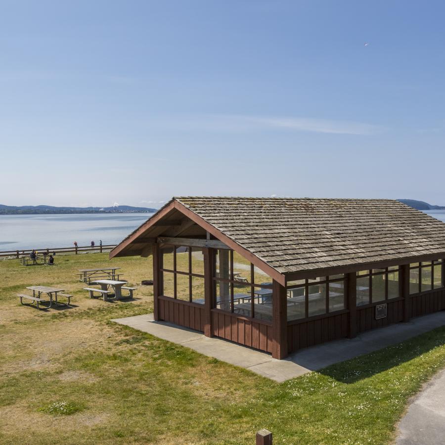 The brown, wooden picnic shelter with a wood shake roof sits in a grassy field, lined with a wood fence. A cement walkway leads down to the shelter, with wooden and cement picnic tables in the grass. Water, land and a blue sky are set in the background. 