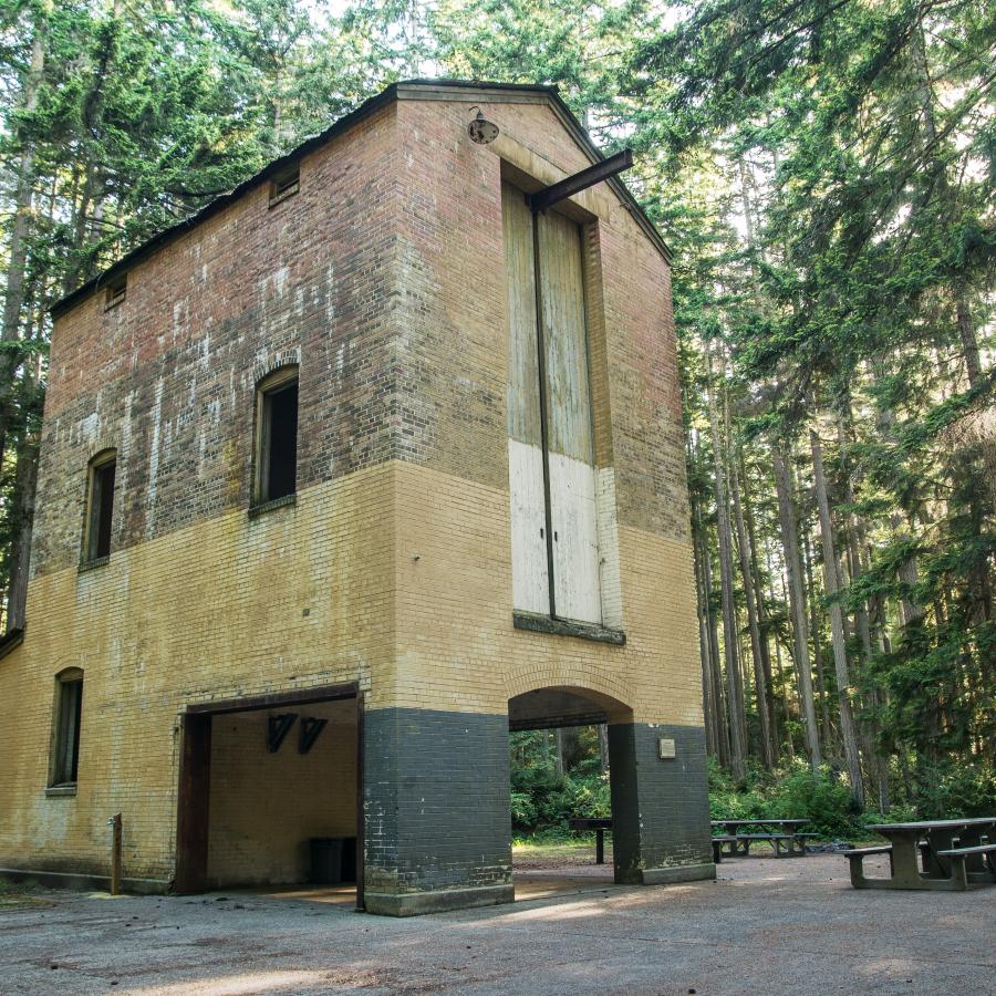 An out of commission xray tower made of brick that's painted black at the bottom, then yellow and plain red brick at the top. Picnic tables and trees surround the tower with sun poking through the trees.