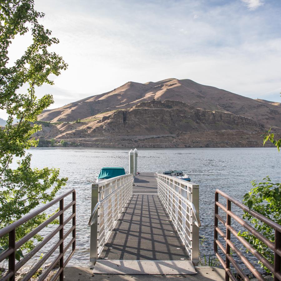 Green leafy trees surround the entrance to the dock ramp with boats tied up to the dock with a brown hillside across the water and a cloudy sky.
