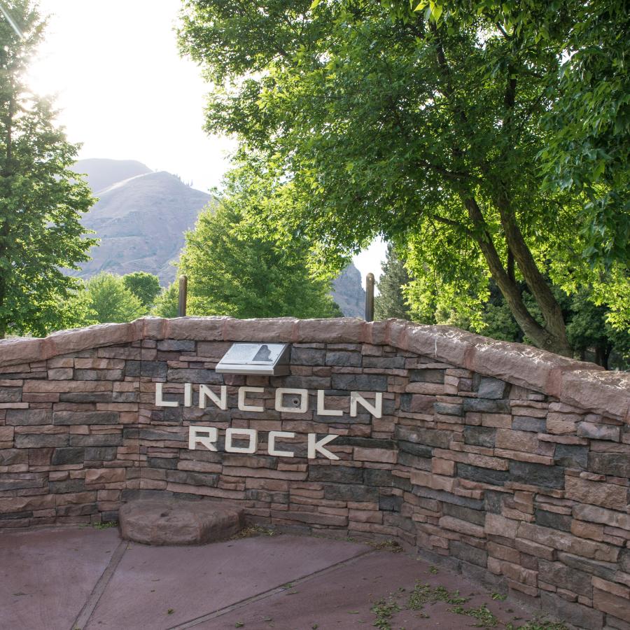 Rock wall structure with 'Lincoln Rock' with tall, leafy trees surrounding the structure. In the distance, a hillside can be seen.