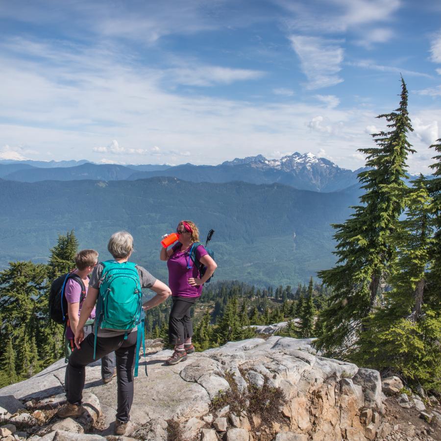 Hikers take a water break on a large boulder with evergreen trees scattered around them. Forested mountains and a snowcapped mountain peak sit in the background with a cloudy blue sky.