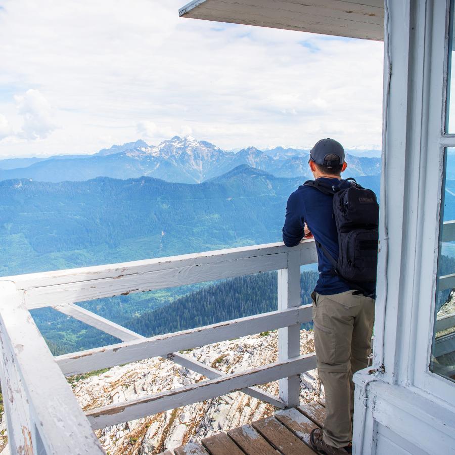 A hiker takes a break while enjoying the view of forested mountains and a snowcapped mountain with a cloudy sky from the lookout tower's deck, with white chipping paint.