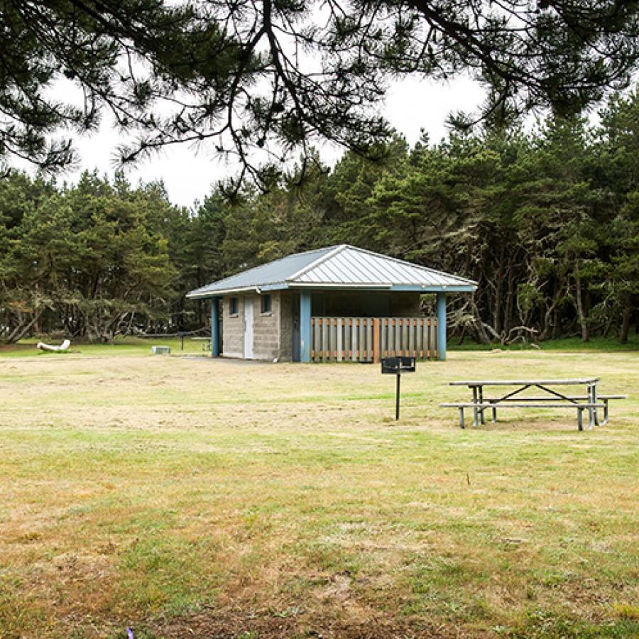 A low restroom building sitting in the middle of a wide green trail. Pines grow in the background