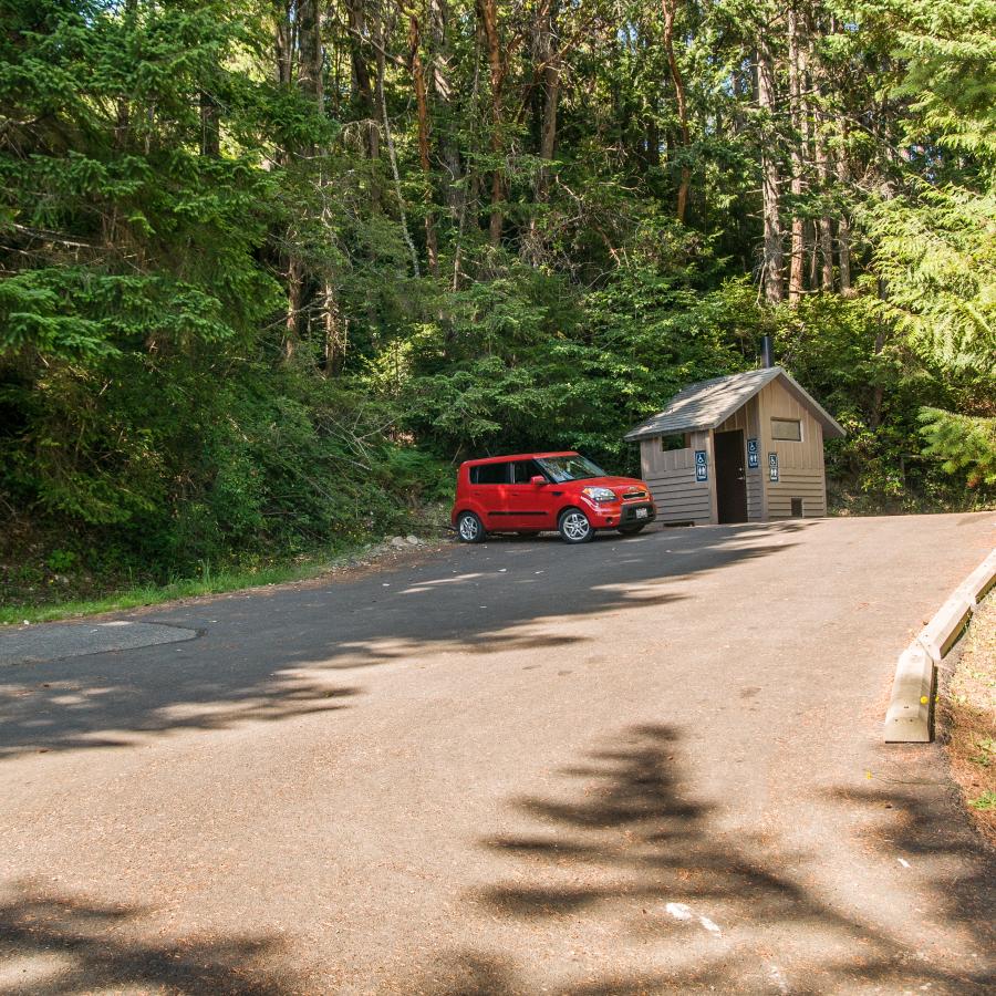 A small red car is parked in an asphalt paved parking lot, lined with cement bumper blocks. A brown restroom sits behind the red car. A sign on the left side of the parking area reads, "No overnight parking or camping" with a Discover Pass required sign underneath. Tall evergreen trees surround the entire area.