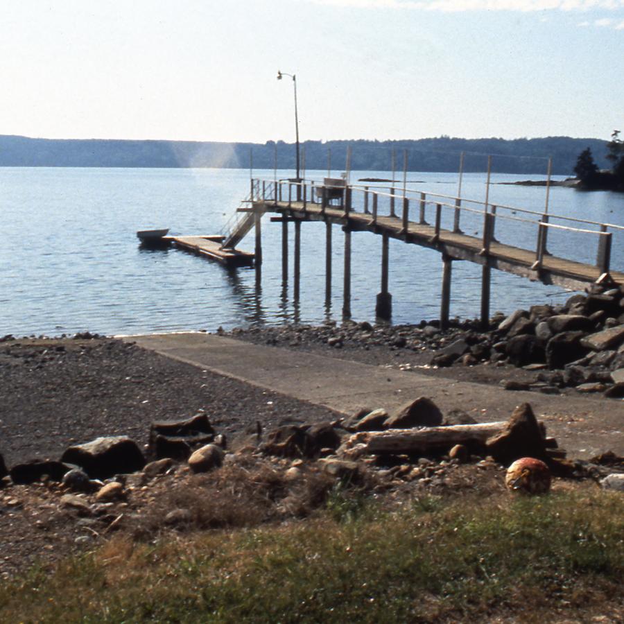 A long pier extends toward a floating boat launch on the water from a rocky shoreline at Triton Cove State Park.