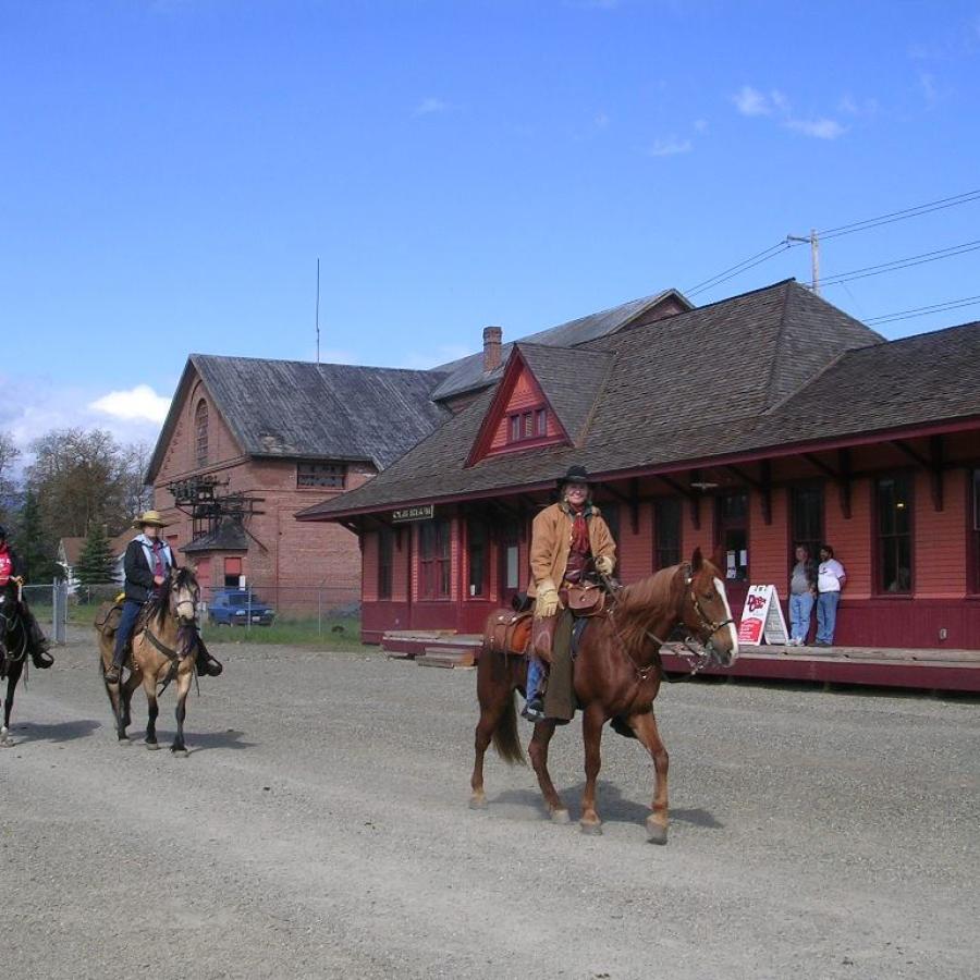 Three horse riders on brown, tan and black horses walk on a dirt road in front of an old trail stations painted orange with red trim. Two people wearing jeans and tshirts lean against the building watching the horse riders go by.