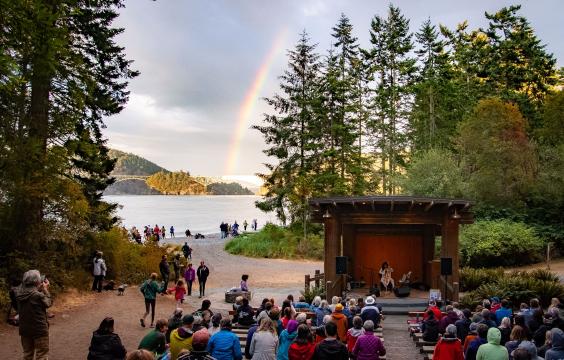 A band playing in an amphitheater at Deception Pass State Park with an audience watching and water, trees and a rainbow behind. 