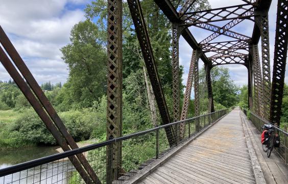 A view looking down an old railroad bridge made of iron on the Willapa Hills State Park Trail. The rails have been replaced with boards and a bicycle is leaning on the rail on the  right side. To the left the Willappa River is seen flowing under the bridge and the  blue sky above is filled with fluffy white clouds.